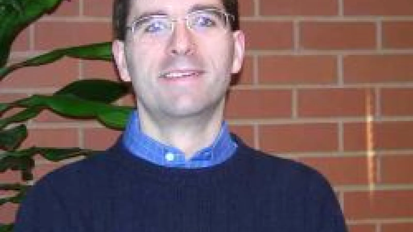 Bradley Ambrose with a brick wall and tall green plant in the background.