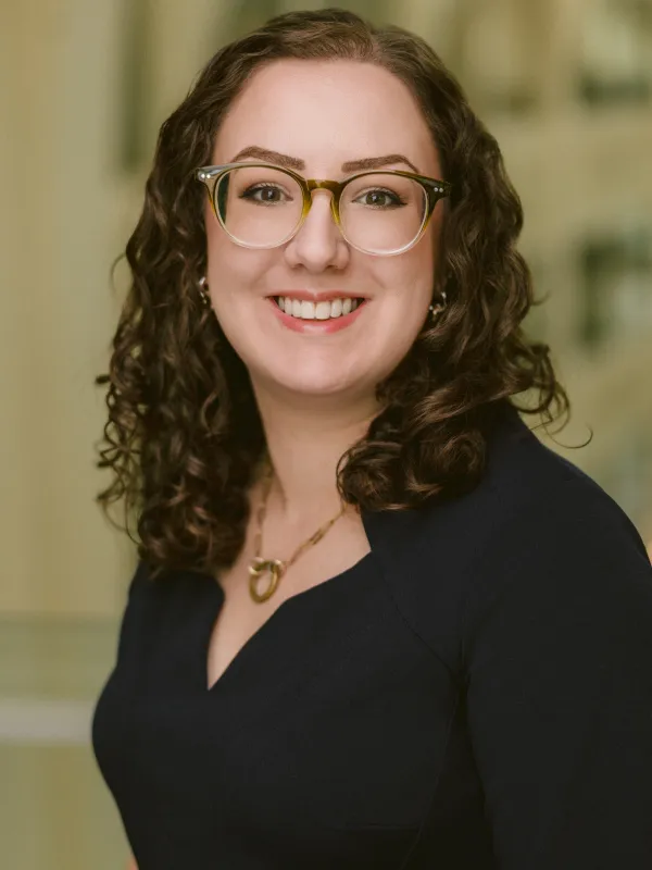 A white woman with brown curly hair, glasses, and a dark shirt on front of a beige blurred background. 