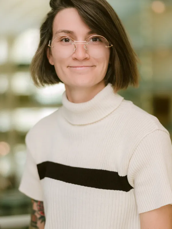 Sierra Brimhall has chin length brown hair, gold glasses, and is wearing a cream turtleneck short-sleeved sweater with a black stripe. She is smiling and the Physics building atrium is in the background behind her. 