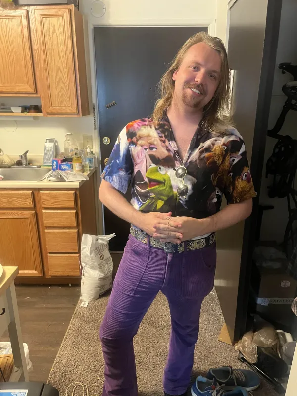 Image of a man in front of a doorway with long blonde hair, purple pants, and a patterned shirt with images of The Muppets